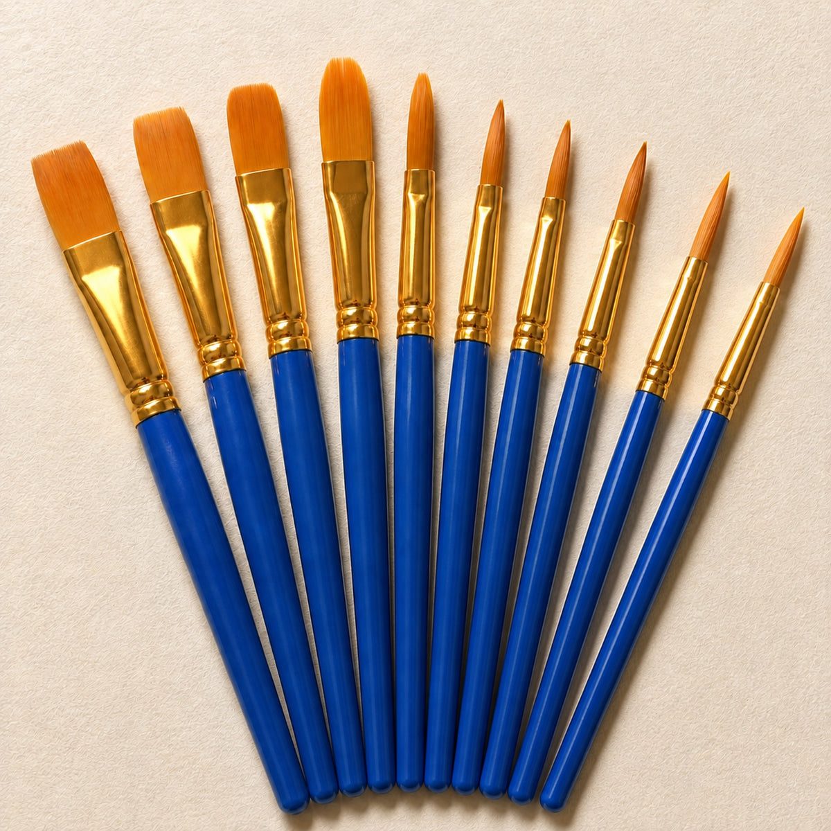 Close view of the ten brushes from one sleeve fanned out on warm ivory, all with blue handles and gold ferrules, in flat, filbert and pointed round shapes.
