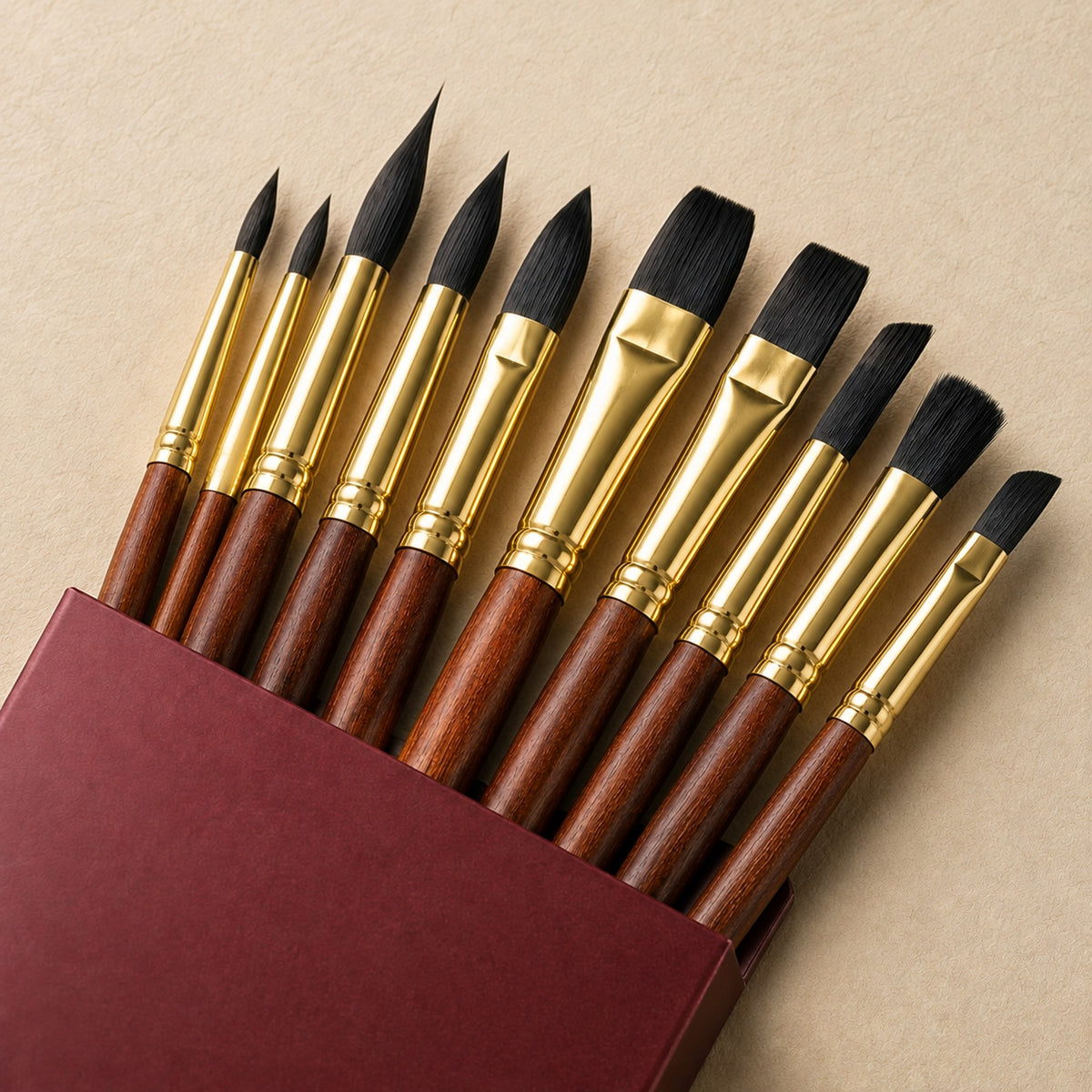 Close view of the ten professional brush heads lifted from the burgundy box, showing pointed rounds, flats, angled shaders and a stippler with polished gold ferrules.