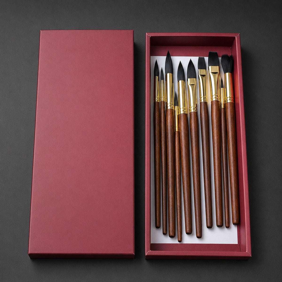 The burgundy presentation box open on a graphite background, holding ten brushes with dark brown wood handles, polished gold ferrules and black filament.